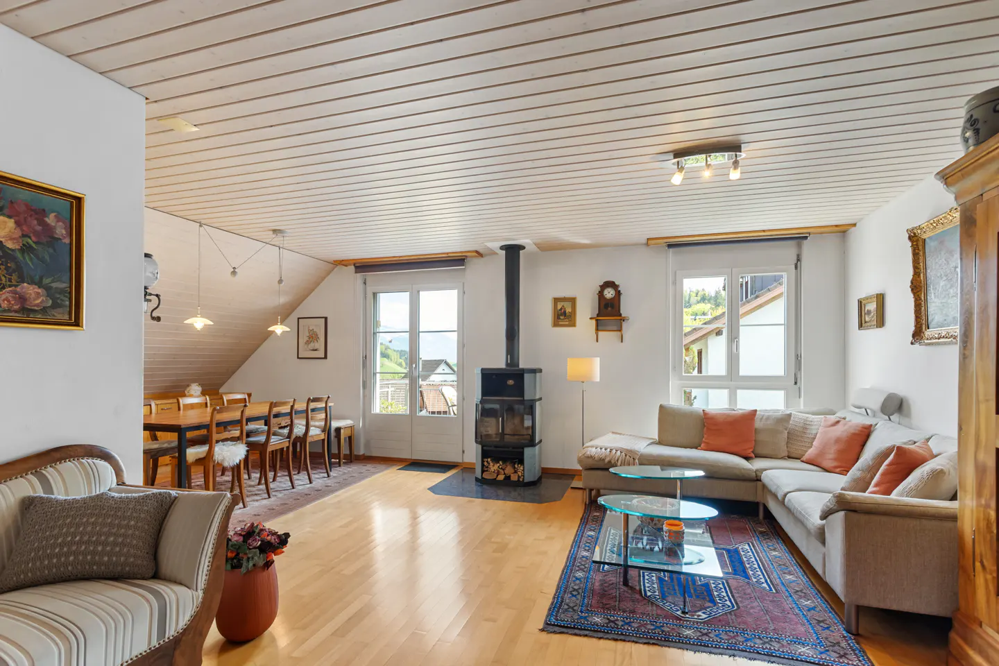 Bright living room with wood floors, white walls, and a wood-burning stove. A beige sectional sofa sits on a patterned rug. Dining table visible in the background.