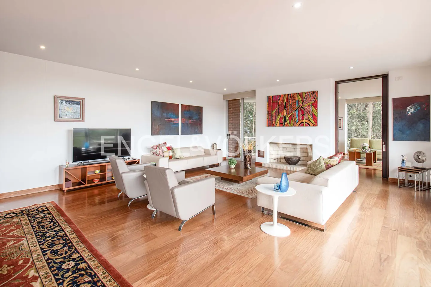 Bright living room with hardwood floors, white sofas, armchairs, and colorful artwork on the walls. A fireplace and TV are also visible.