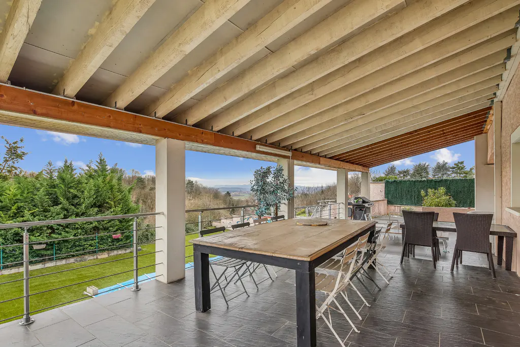 Covered patio with a large wooden table, chairs, and a view of green trees and a blue sky.