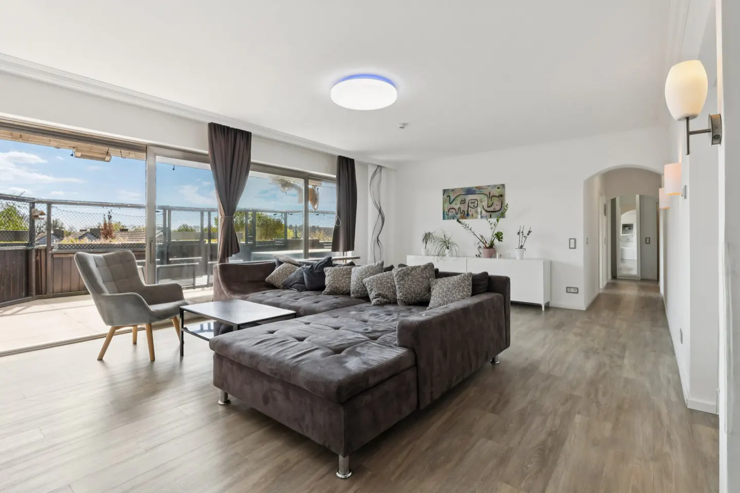 Bright living room with a gray sectional sofa, chair, and wood floors. Large windows lead to a balcony with a view.