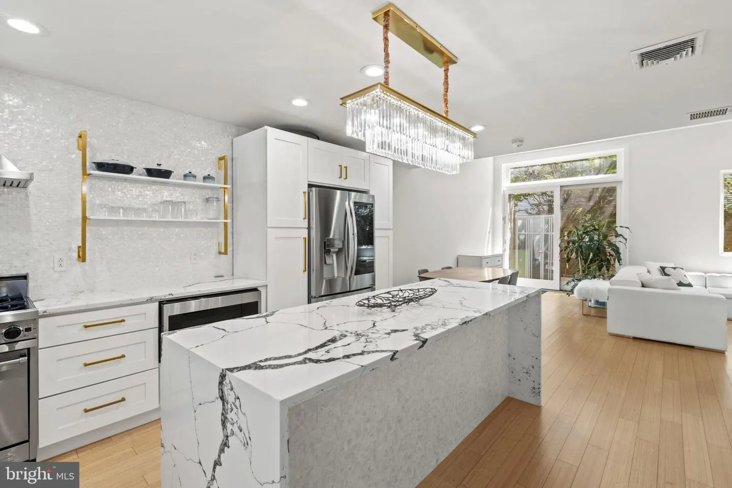 A bright, modern kitchen with white cabinets, marble countertops, and a crystal chandelier. A stainless steel refrigerator is visible.
