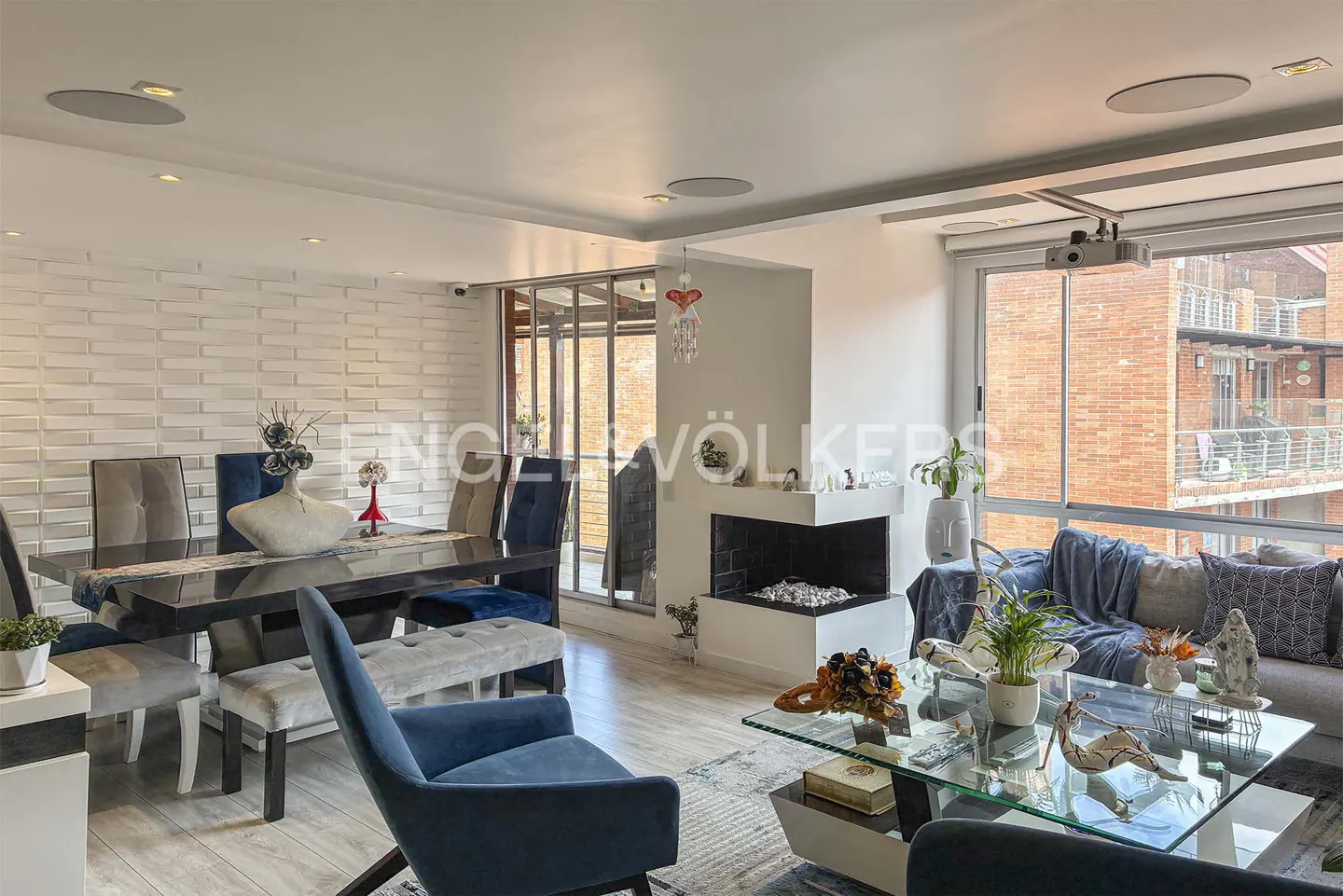 Modern open-plan living area with a dark dining table, blue velvet chairs, a glass coffee table, and a corner fireplace.