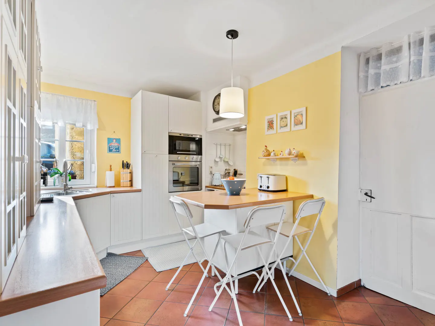 Bright kitchen with yellow walls, white cabinets, and terracotta tile floor. A small table with folding chairs sits in the center.