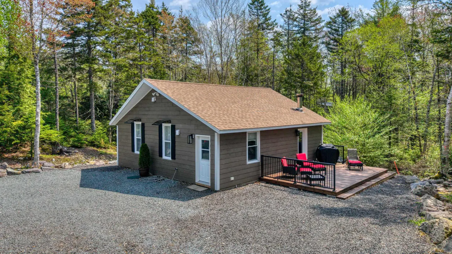 A cozy, one-story brown house with a deck and red chairs, surrounded by a lush green forest.