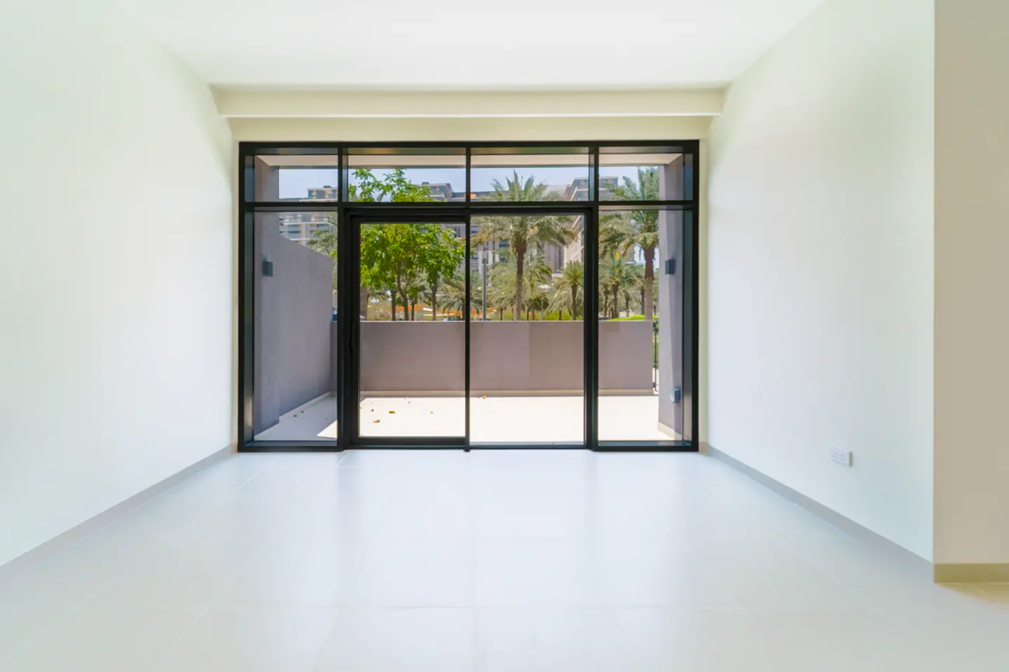 Empty room with white walls and tile floor. Large black-framed sliding glass doors lead to a balcony with palm tree views.