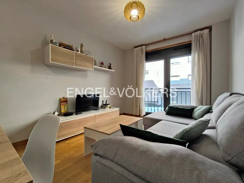 Living room with gray sectional sofa, wood floors, and a TV on a light wood console. A balcony is visible through the glass doors.