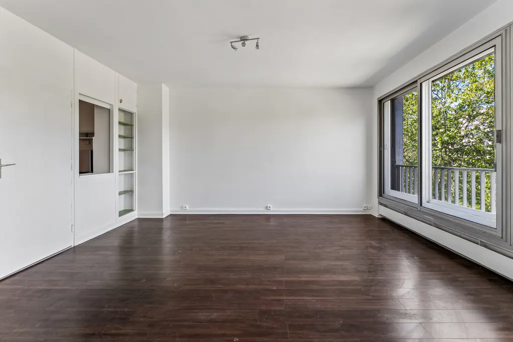 Bright, empty room with dark wood floors, white walls, and large windows showing trees and a balcony. Built-in white cabinets on the left.