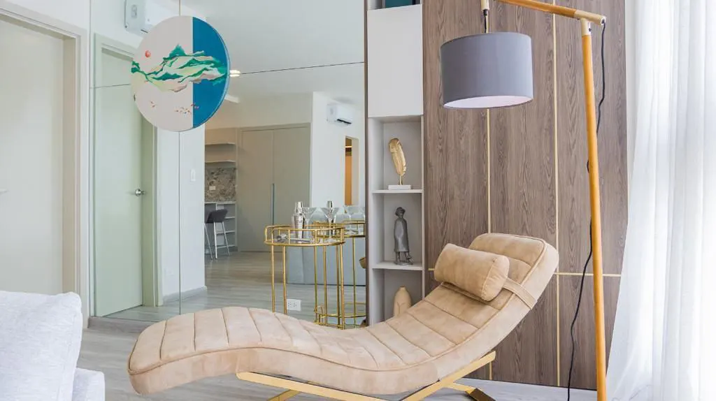 A modern living room with a beige chaise lounge, a wooden floor lamp with a gray shade, and a mirrored wall with art.