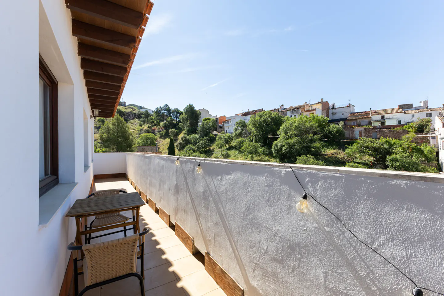Balcony view with a small table and chairs, white wall, string lights, and a view of houses and trees on a hill.