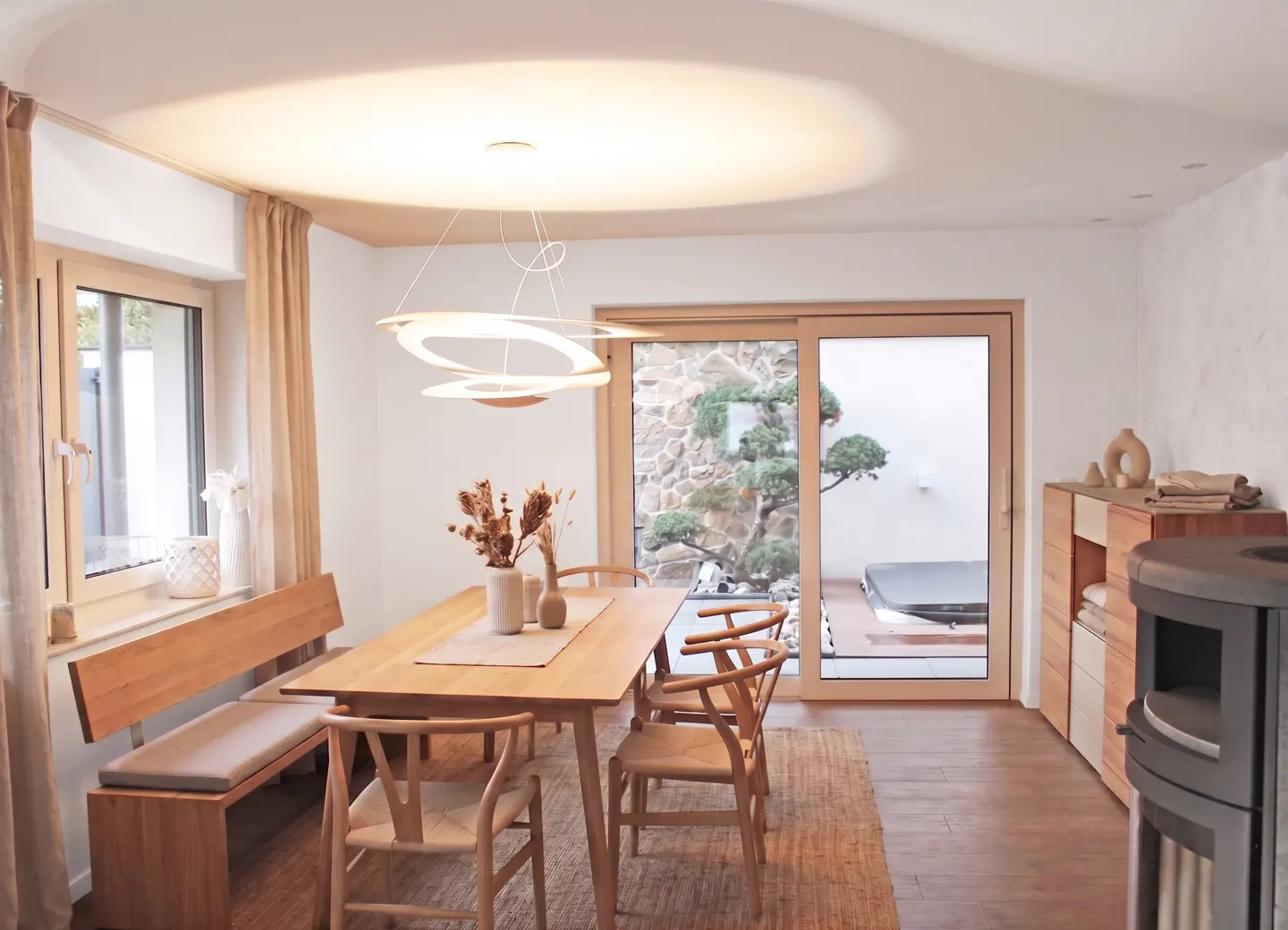 Bright dining room with a wooden table, chairs, and bench. A modern chandelier hangs above. Sliding glass doors lead to a patio with a tree and hot tub.