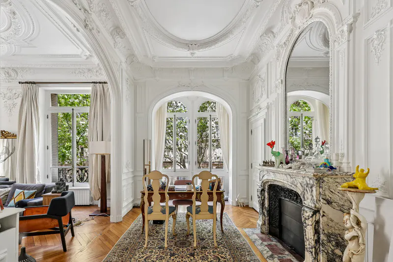 Elegant room with white ornate molding, a marble fireplace, a desk with two chairs, and large windows overlooking trees.