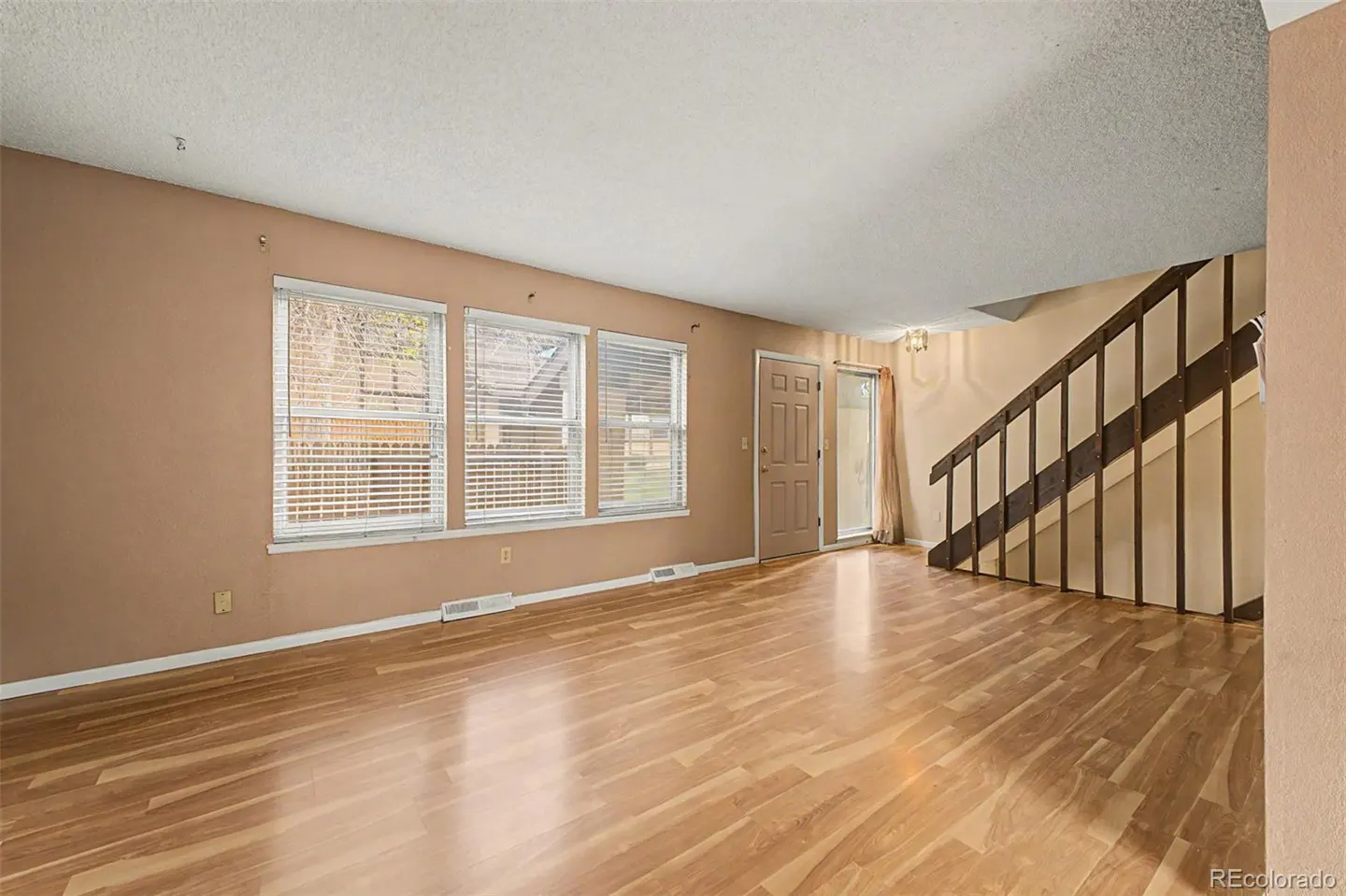 A living room with wood floors, tan walls, three windows with blinds, and a staircase.