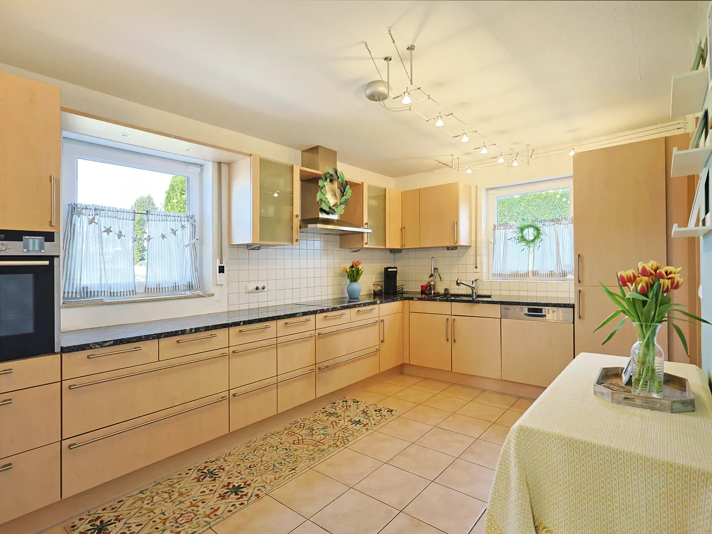 Bright kitchen with light wood cabinets, black countertops, and tile floors. A table with tulips sits in the foreground.