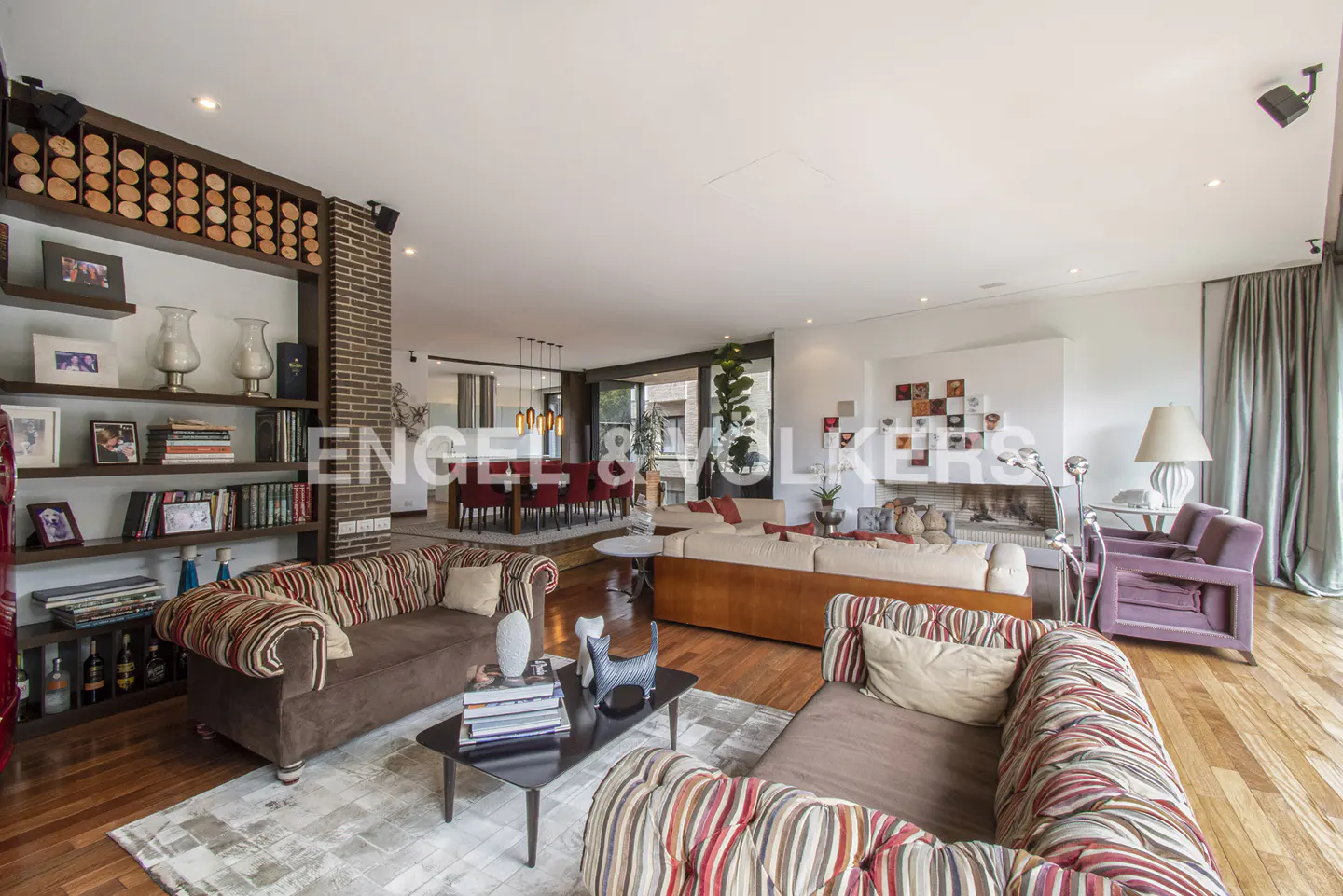 Open-concept living room with hardwood floors, striped sofas, and a dining area in the background. A bookshelf and fireplace add character.