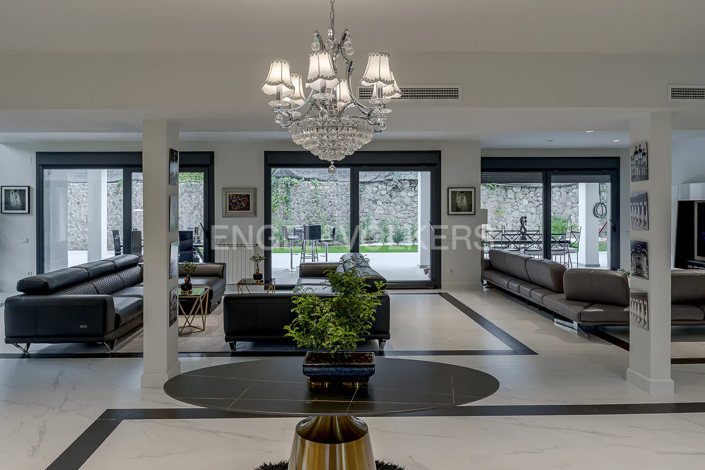 A modern living room with black sofas, marble floors, and a crystal chandelier. Sliding glass doors lead to an outdoor area.