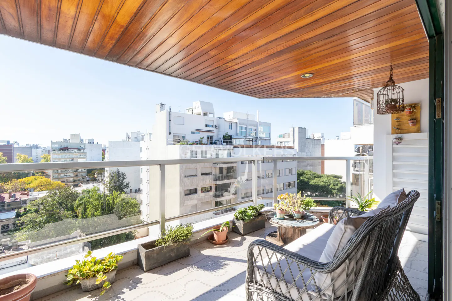 A sunny balcony with wicker chairs, potted plants, and a city view. The ceiling is wood paneled.
