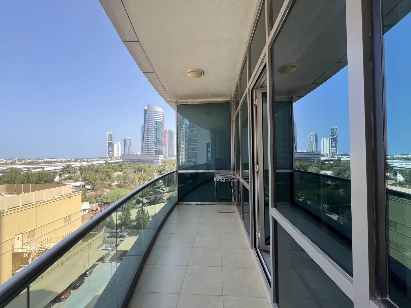 Balcony view of a city skyline. Glass railings and sliding doors frame the cityscape. A clothes drying rack stands on the tiled floor.