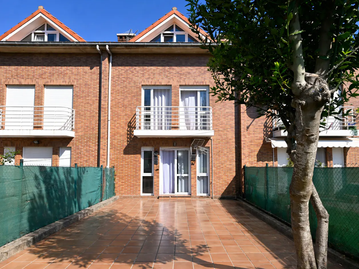 Brick townhouse with a tiled patio, green fence, and a tree on the right. Balconies with white railings are on the second floor.