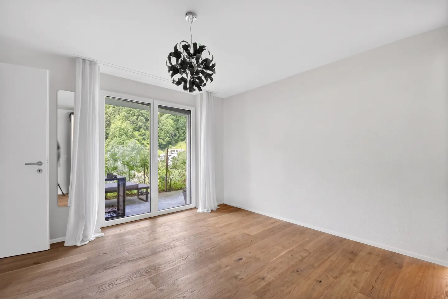 Bright, empty room with wood floors, white walls, and a modern black chandelier. Sliding glass doors lead to a patio with a table and green trees.