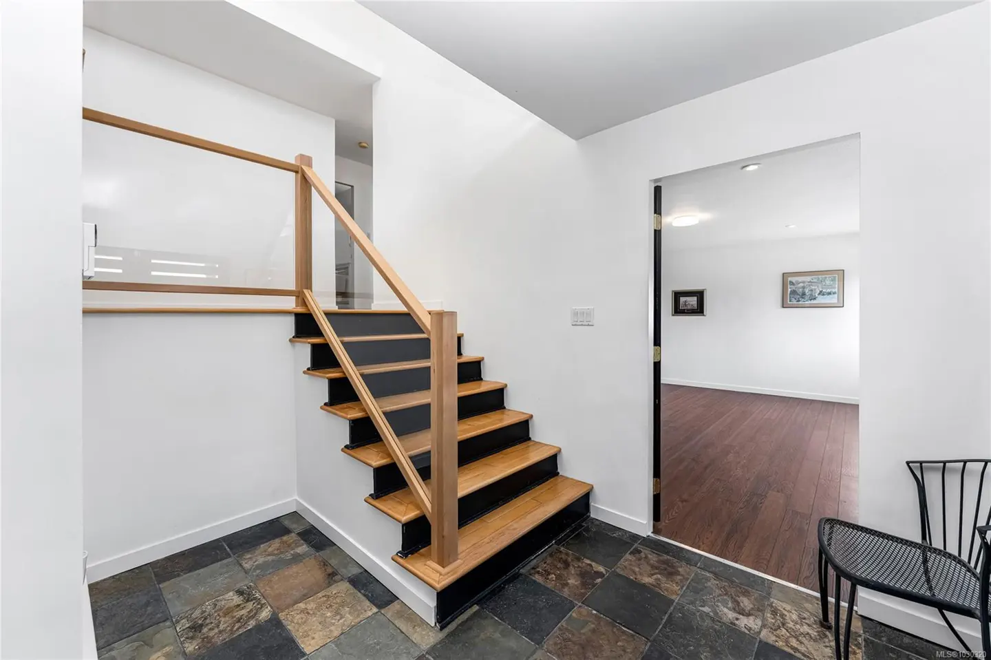 Foyer with wood and black stairs, glass railing, and slate tile floor. A doorway leads to a room with hardwood floors.