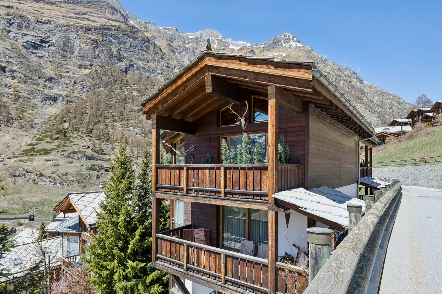 Two-story wooden chalet with balconies, set against a backdrop of mountains and a clear blue sky.