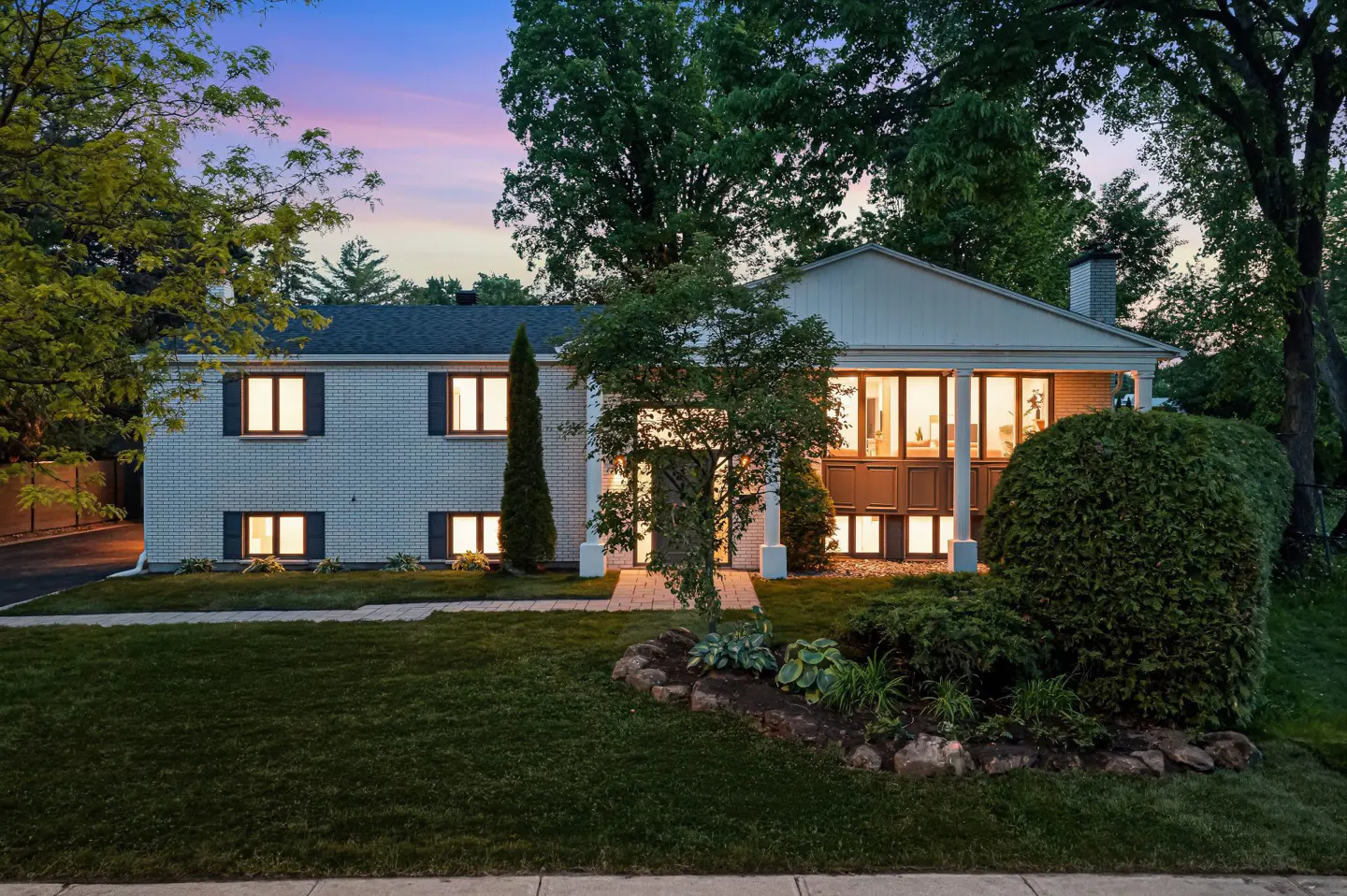 White brick split-level home at dusk with illuminated windows, lush landscaping, and a sunset sky in the background.