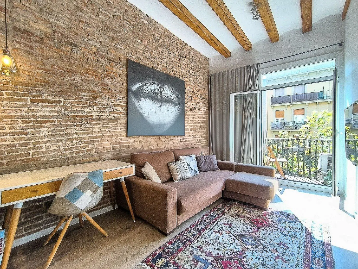 Living room with exposed brick wall, wooden beams, and balcony. Brown sectional sofa, desk, and patterned rug. Large lip art on the wall.