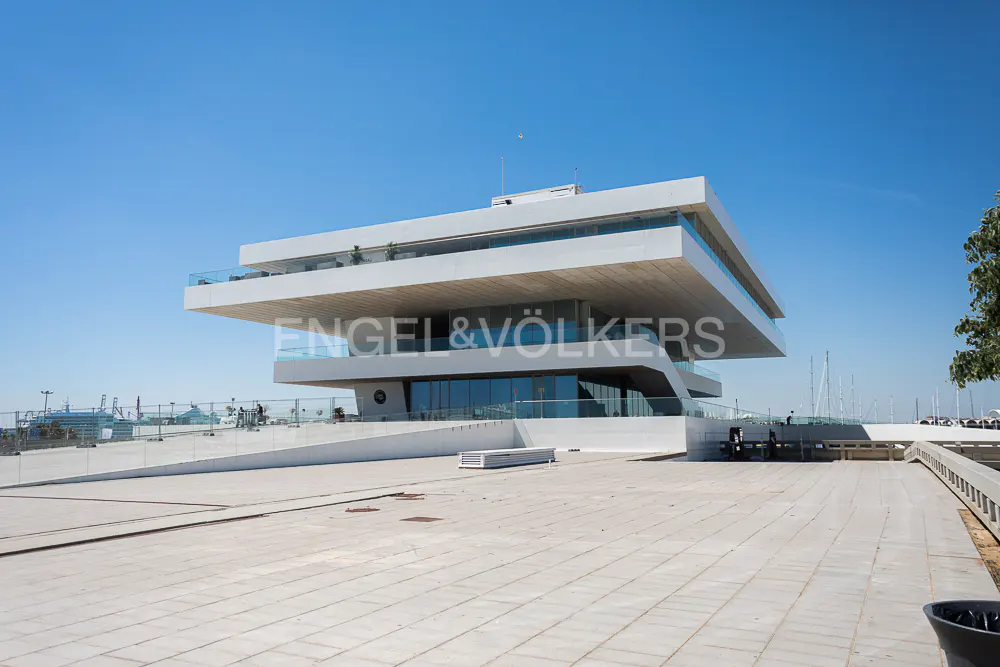 Modern white building with glass railings under a clear blue sky. The building has the Engel & Völkers logo. A paved area is in the foreground.