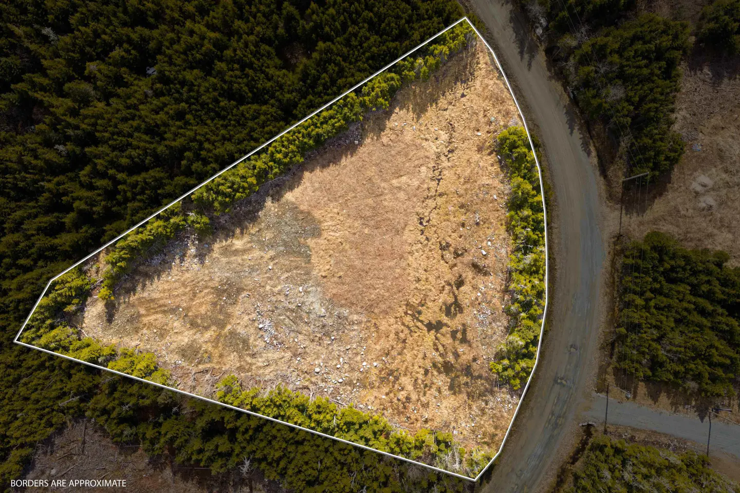 Aerial view of a vacant lot with a white border, surrounded by trees and a road. The lot is mostly brown with some green vegetation.