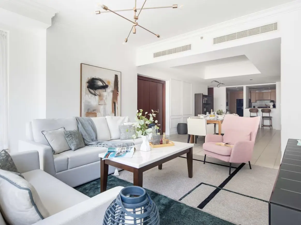 Bright, open-plan living room with white sofas, a marble-topped coffee table, and a pink armchair. A modern chandelier hangs above.