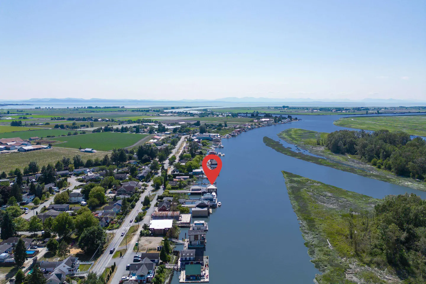 Aerial view of houseboats on a river, marked with a red location pin. Green fields and mountains are in the background.