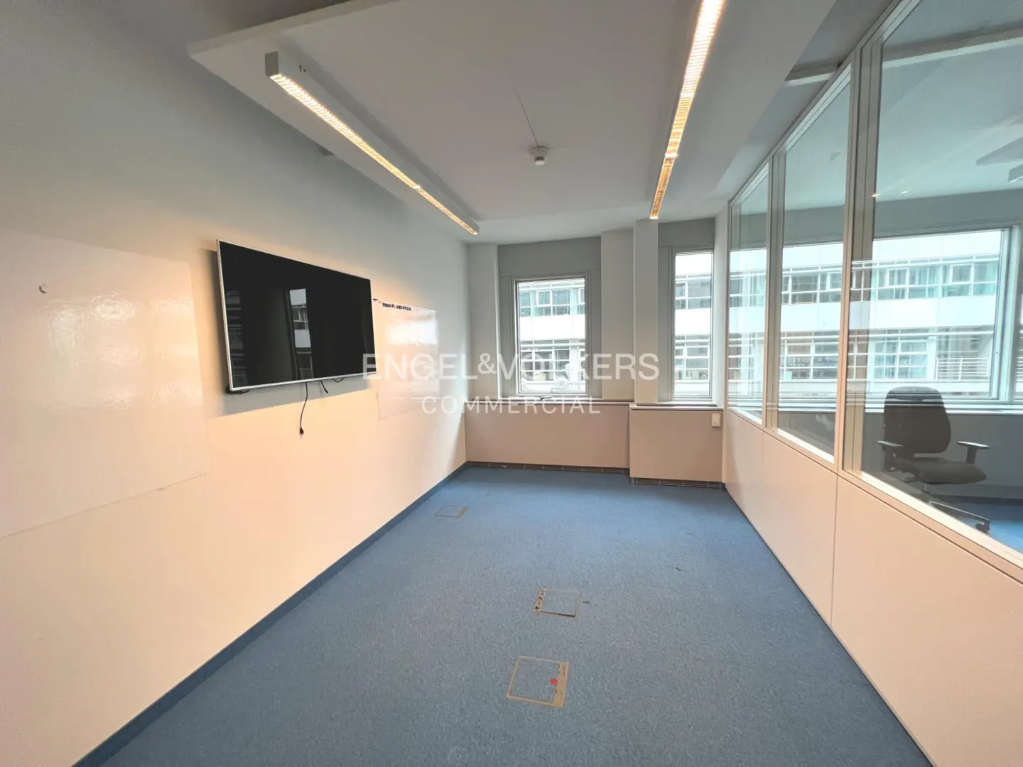 Empty office space with blue carpet, white walls, and a large TV. Windows offer a city view. "Engel & Volkers Commercial" logo is visible.