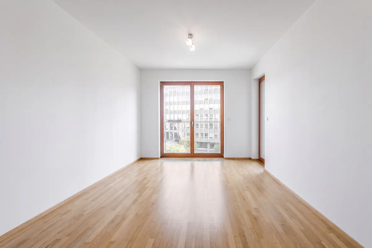 Bright, empty room with hardwood floors, white walls, and a glass door to a balcony with a city view.