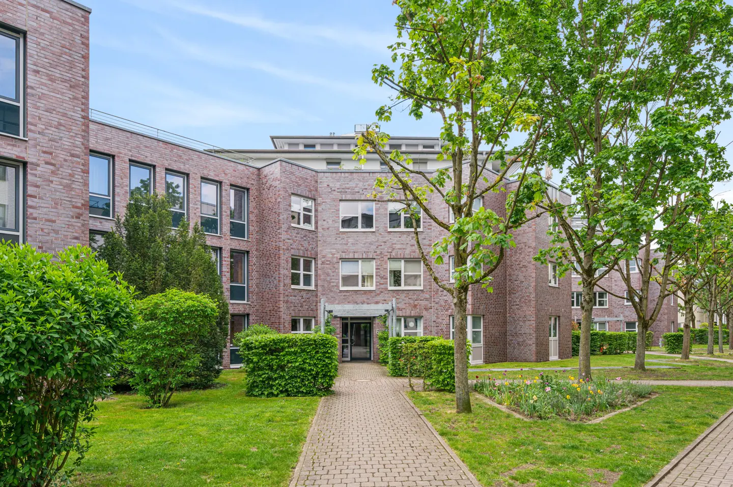 A modern multi-story brick apartment building with a paved walkway, green lawns, and lush trees under a blue sky.