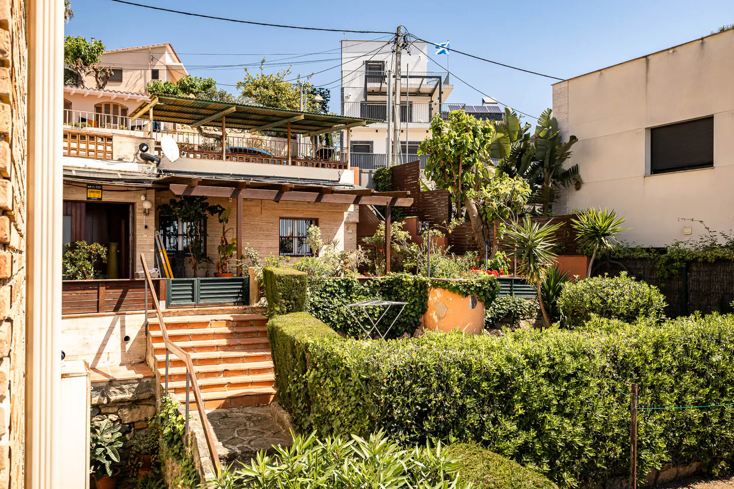 Exterior view of a multi-level home with a garden. Brick stairs lead to a patio with a pergola. Green hedges and plants surround the property.