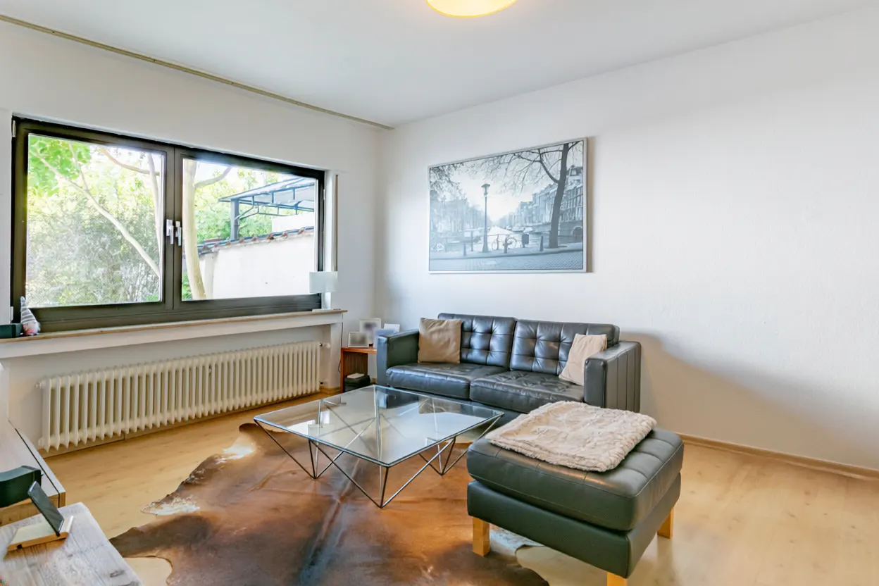 Bright living room with a black leather sofa, ottoman, and a glass coffee table on a cowhide rug. A large window and a city print add character.