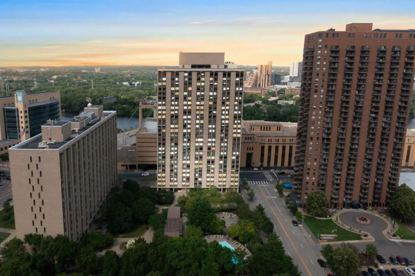 Aerial view of tall apartment buildings with green trees, a pool, and a city skyline at sunset.
