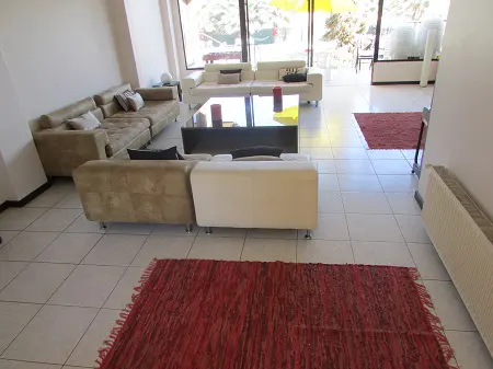 Living room with tile floors, beige and white sofas, a black coffee table, and red rugs. Large windows let in natural light.