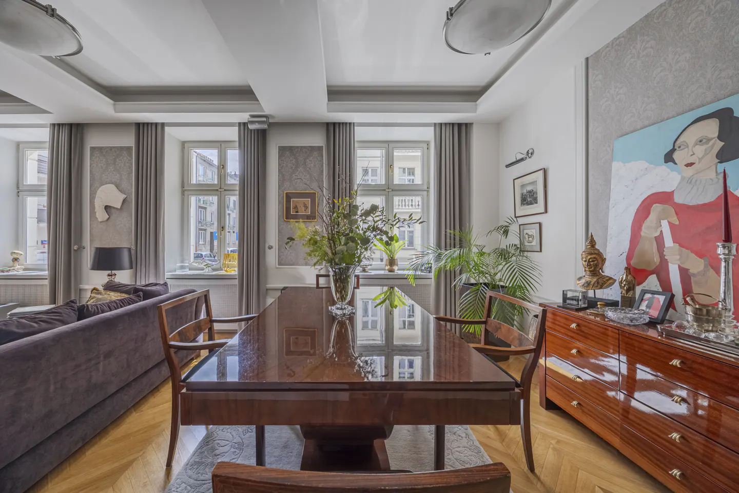 A bright living room with a long, dark wood table, chairs, and a brown sofa. Windows with gray curtains line the walls. A painting hangs above a wood dresser.