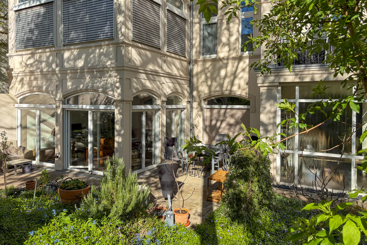 Exterior view of a beige two-story house with a patio, plants, and outdoor furniture. Sunlight filters through the trees, casting shadows on the building.