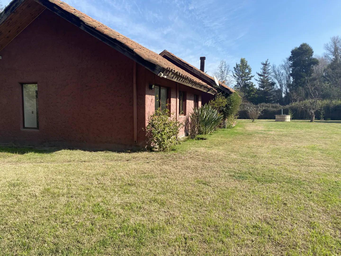 A rustic, reddish-brown house with a tiled roof sits on a large, grassy lawn under a clear blue sky.