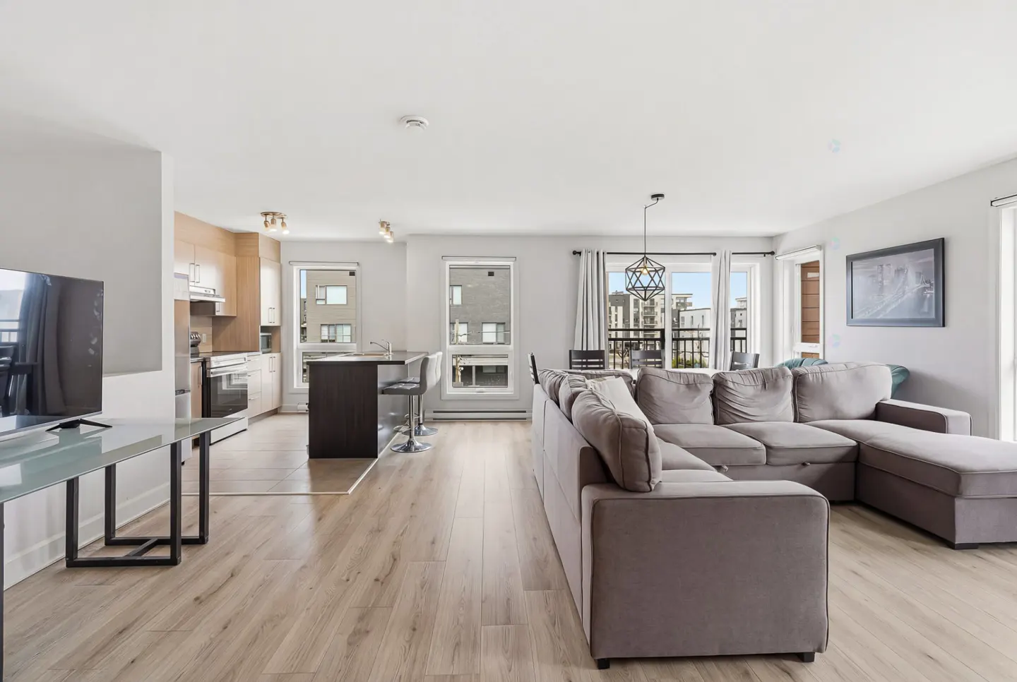 Bright, open-concept apartment with a gray sectional sofa, wood floors, and a kitchen with a breakfast bar. Windows offer city views.