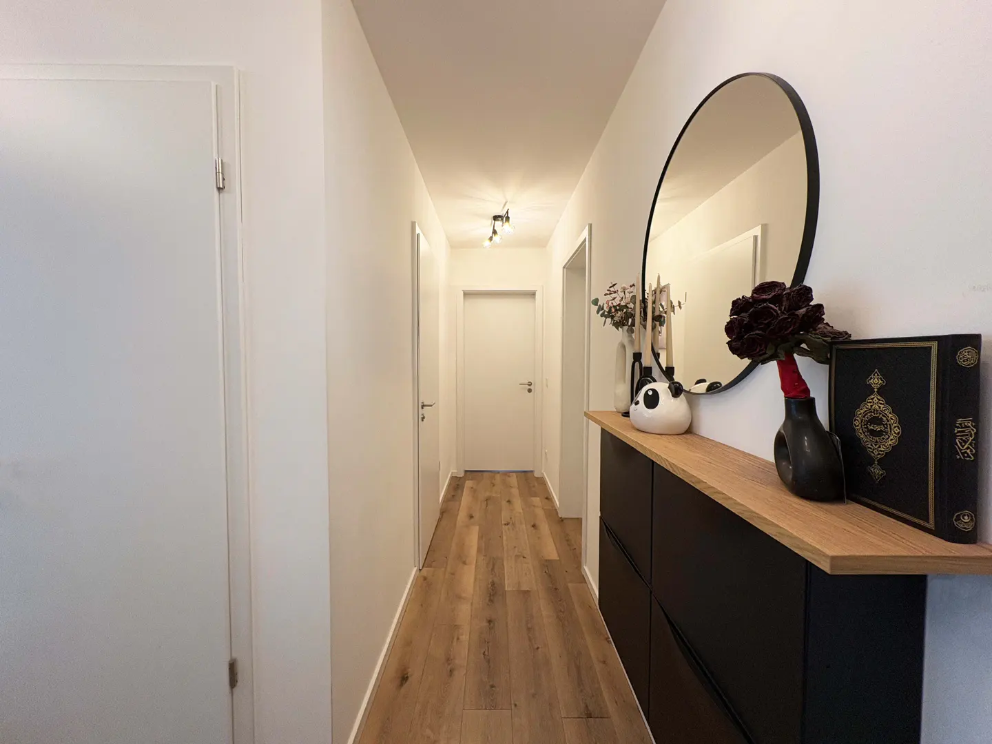 Hallway with wood floors, white walls, and a black cabinet with a wood top. A round mirror hangs above the cabinet.