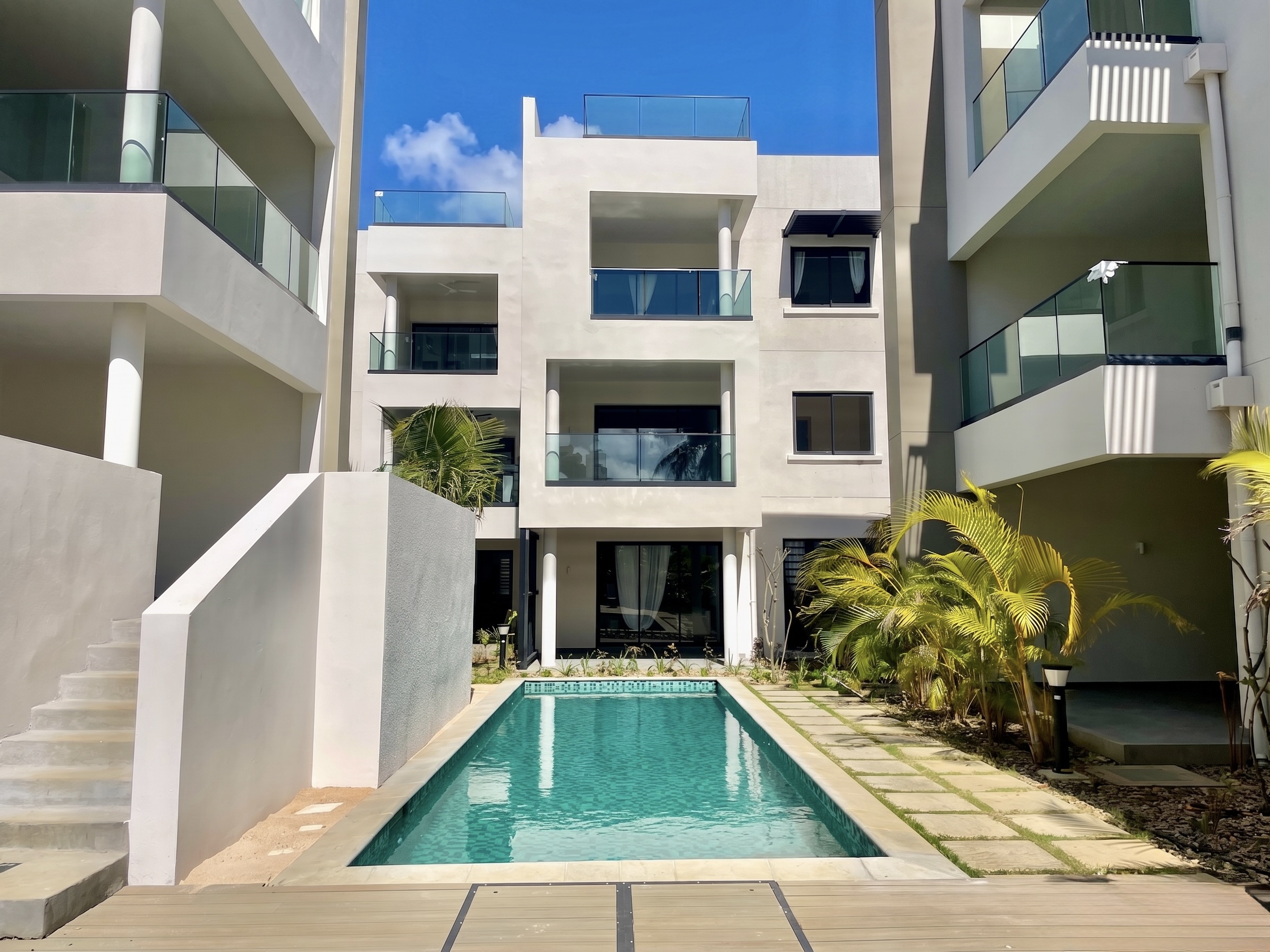 Modern white apartment complex with a rectangular swimming pool, glass balconies, and tropical landscaping under blue sky. [AI-generated text]