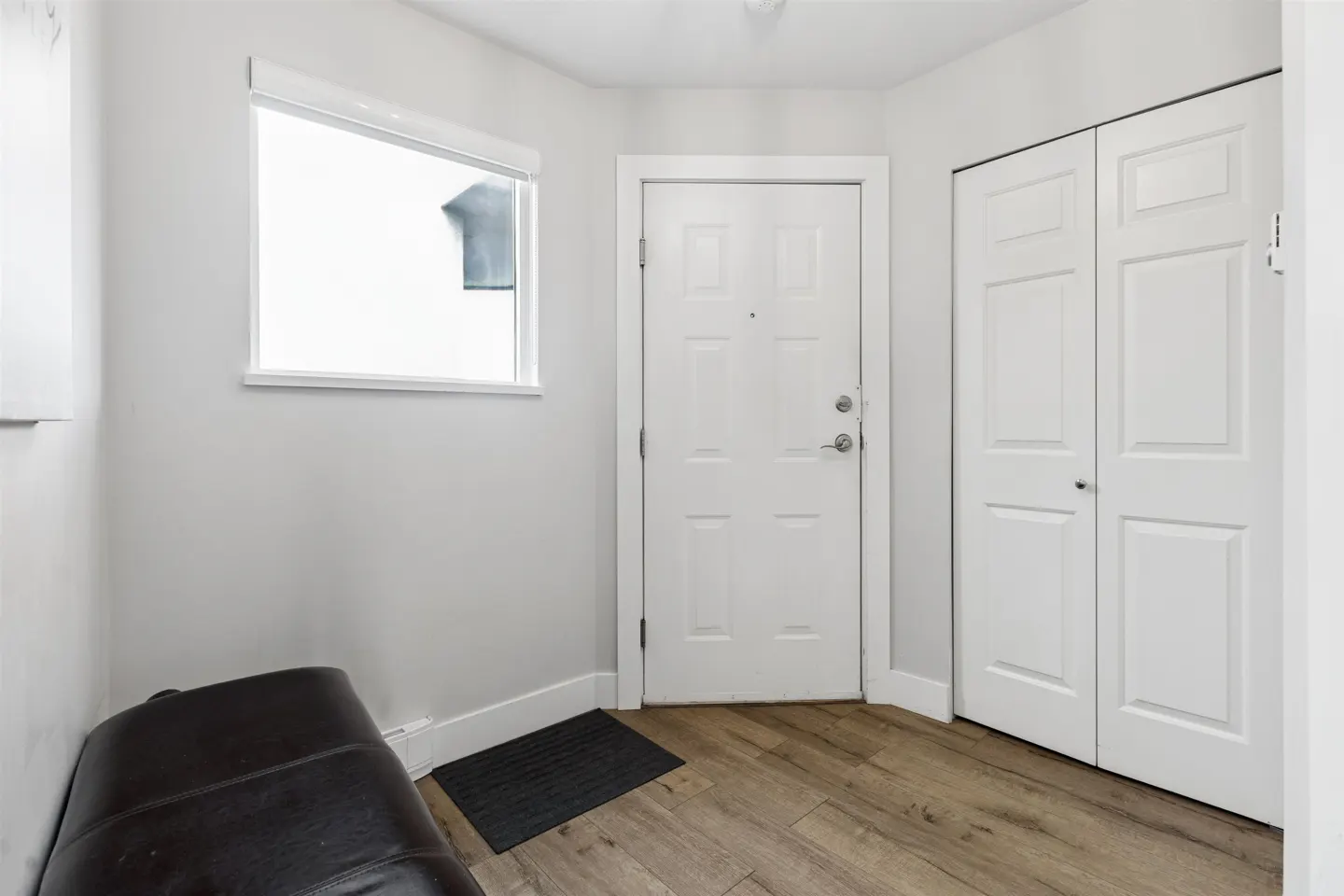 Entryway with light wood floors, white walls, and a white front door. A dark brown bench sits near a window. A black doormat is on the floor.