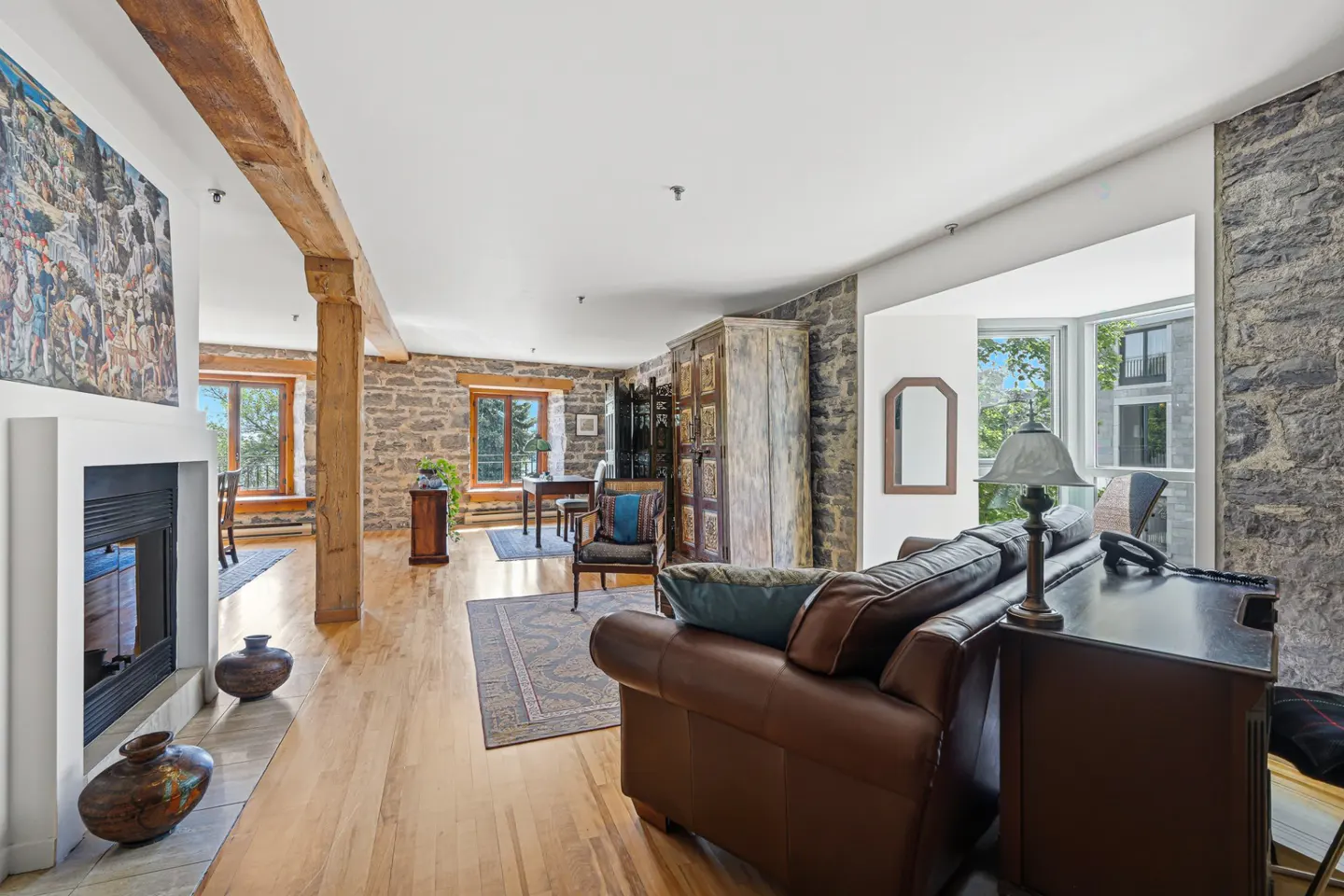 A living room with stone walls, hardwood floors, and a brown leather sofa. A fireplace, wooden beams, and a dining area are also visible.