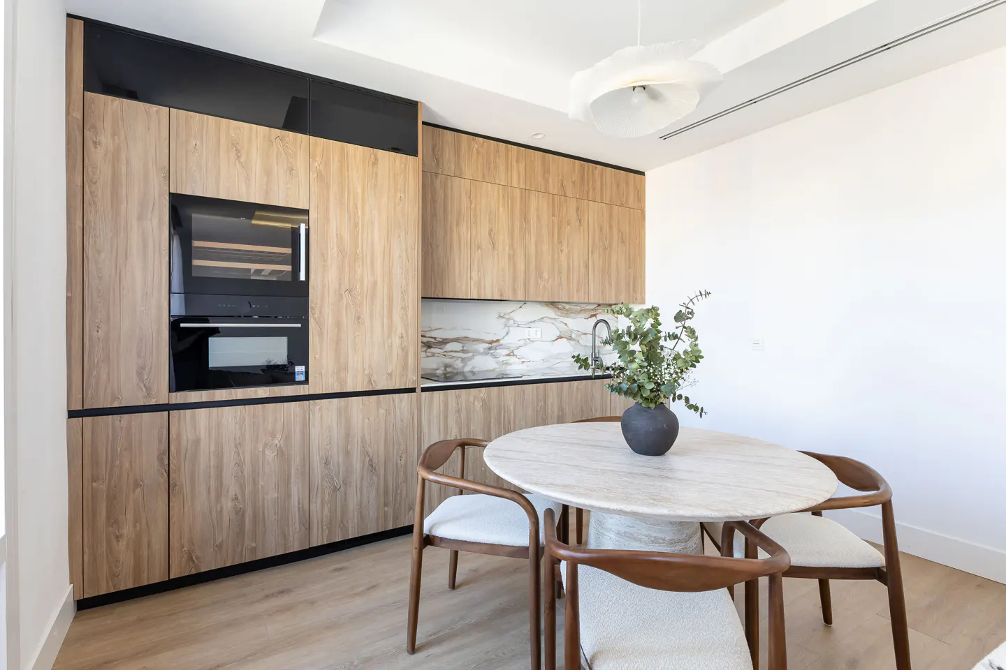 A modern kitchen with wood cabinets, black accents, and a marble-topped round table with four wooden chairs. A vase of greenery sits on the table.