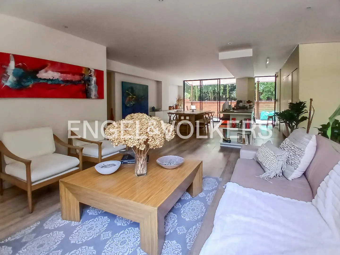 Open-concept living room with wood floors, a large coffee table with flowers, and a dining area visible in the background.