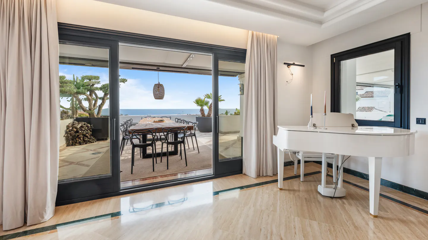 A bright room with a white piano, marble floors, and a sliding glass door to a patio with an ocean view.