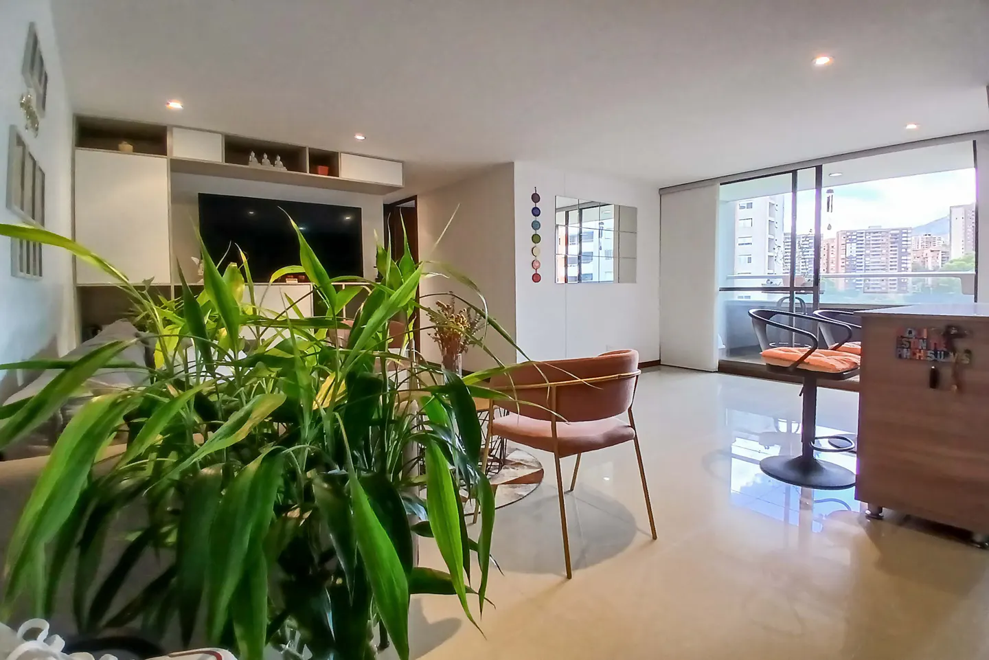 Modern living room with a large green plant in the foreground, a pink accent chair, and a balcony with city views.